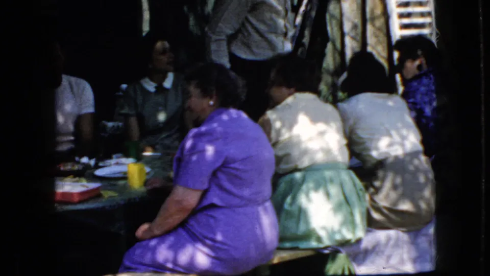 California 1969 home-movie frame showing adult women and other attendees seated at an outdoor picnic table in mottled sunlight.