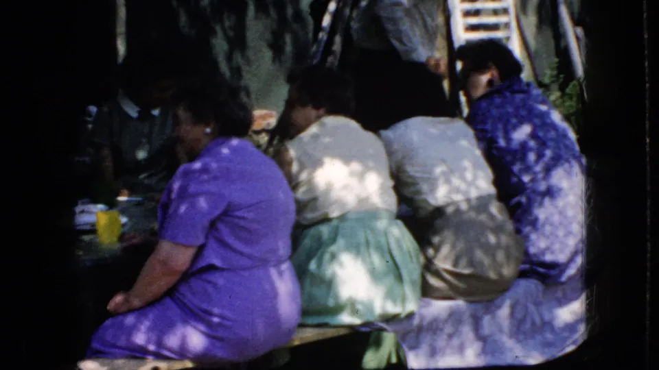 California 1969 home-movie frame with adult women in colorful dresses seated together at a shaded church picnic table.