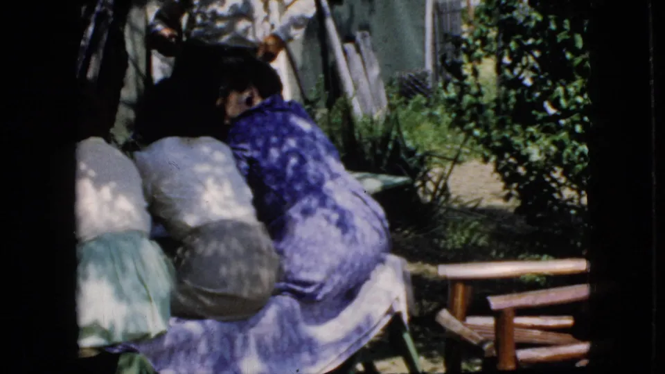 California 1969 home-movie frame with adult women seated at the edge of a shaded picnic table, with greenery and a wooden lawn chair nearby.