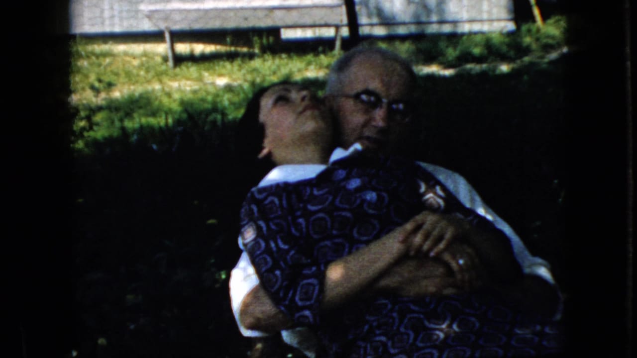 Elderly man in glasses holds a woman in a blue patterned dress on a shaded lawn.