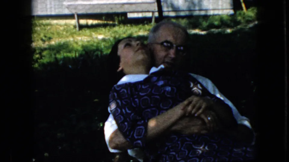 In 1969 California home-movie footage, an elderly man in glasses and a white shirt embraces a dark-haired woman in a blue patterned dress outdoors on grass.