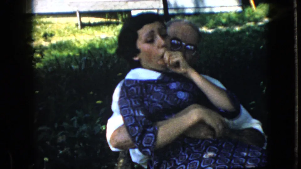 The woman appears to cough or laugh while being held by the elderly man, with grass and sunlit background visible in the 1969 California home-movie frame.