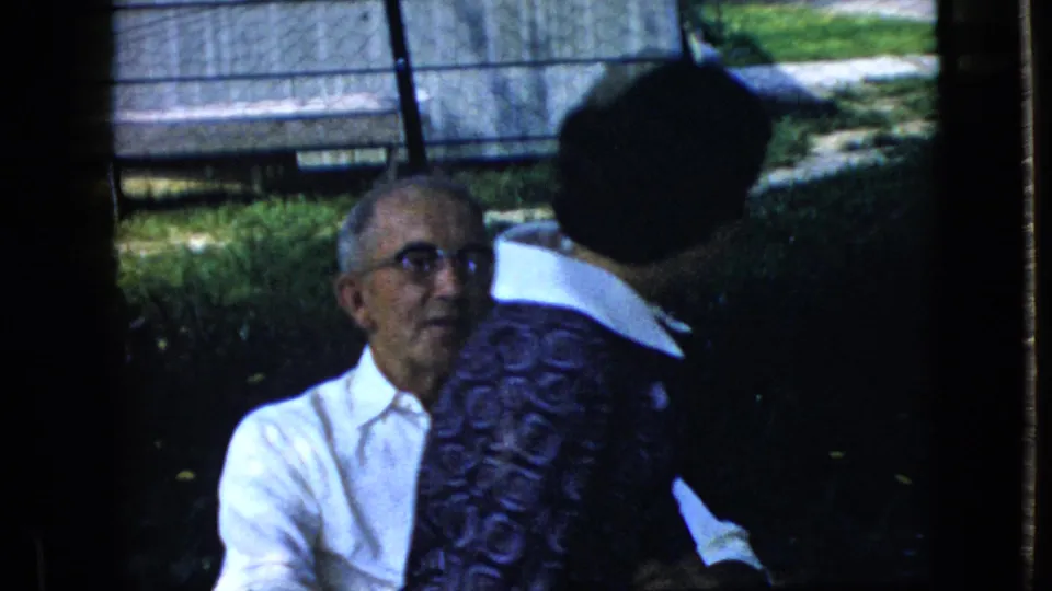 The elderly man in glasses looks toward the camera as the woman turns away, ending the brief outdoor embrace in the 1969 California 8mm footage.
