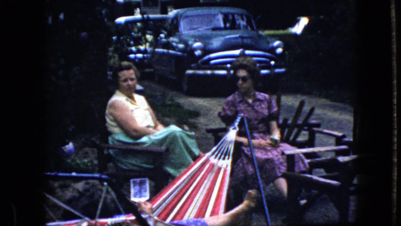 Two women sit outdoors beside a striped hammock with an older dark sedan behind them.