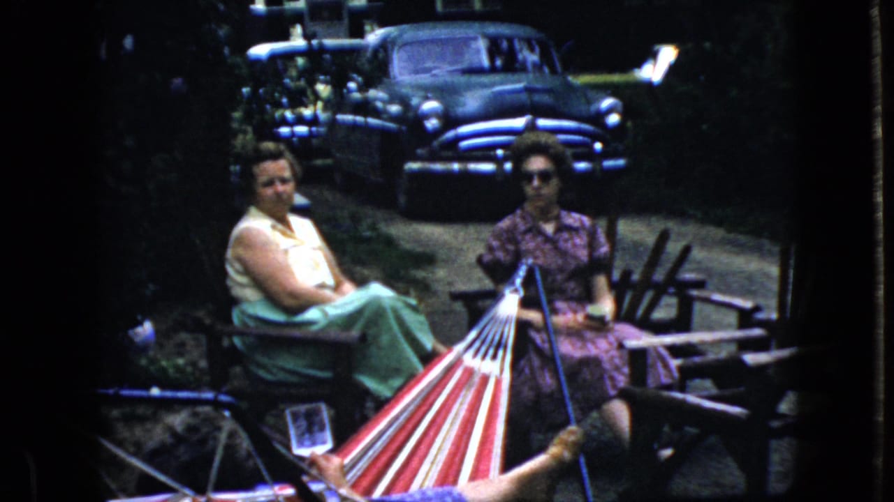 Two women sit near a striped hammock while a dark sedan is parked in the background.