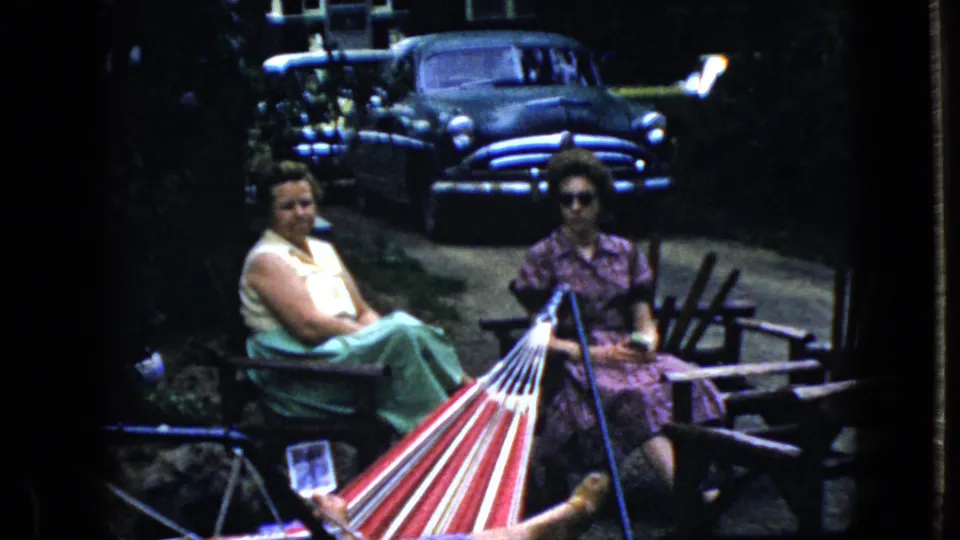 Two women sit facing the camera in a shaded California yard or driveway scene from 1969, with a striped hammock crossing the foreground and a dark older sedan visible behind them.
