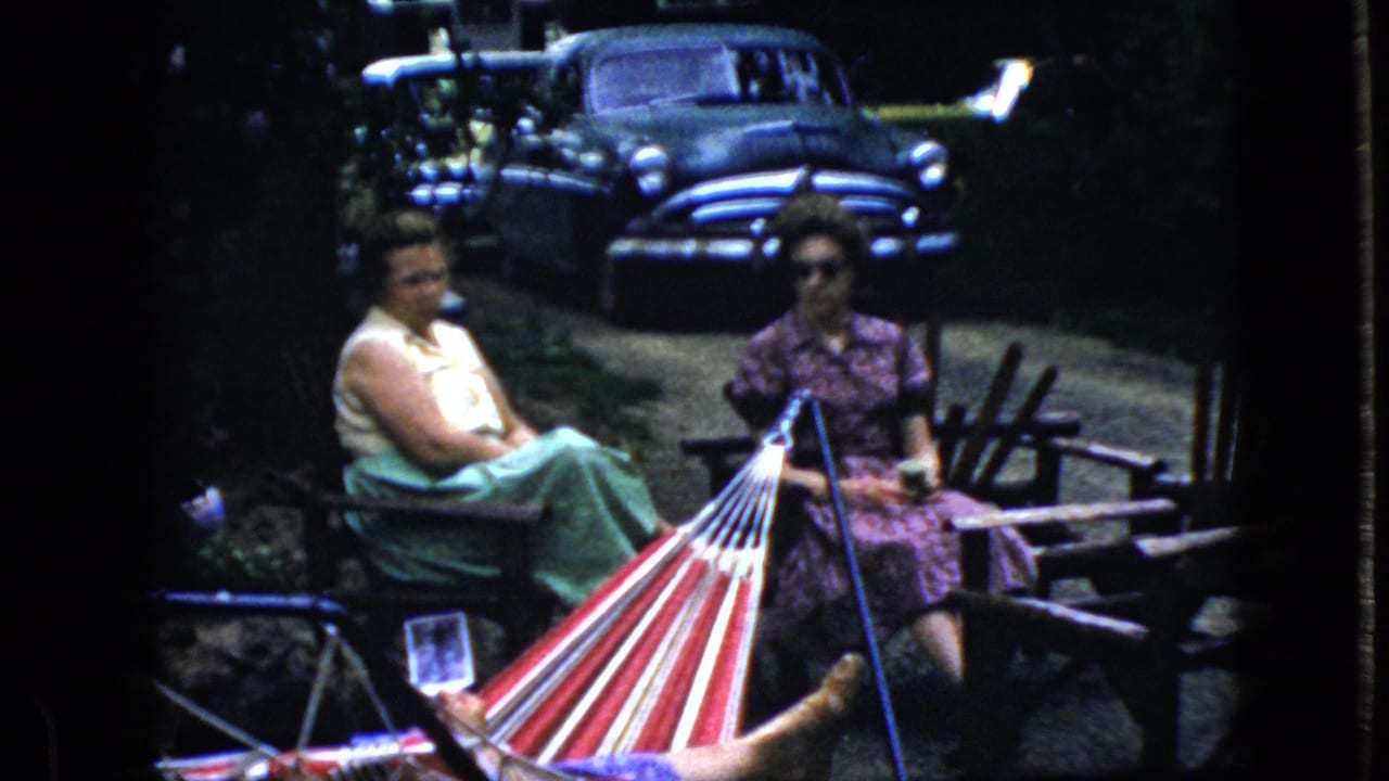 Two women sit outdoors near a striped hammock with a dark sedan parked behind them.