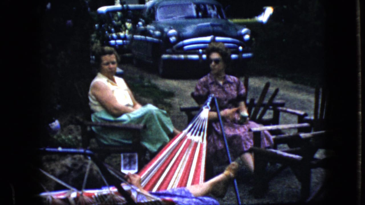 Two women sit near a striped hammock while an older dark car is parked in the background.