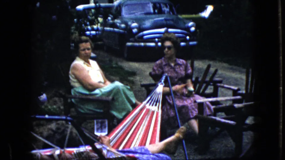 The frame shows two women in outdoor seating near a striped hammock, with a dark mid-century sedan in the shaded background of the California home-movie scene.