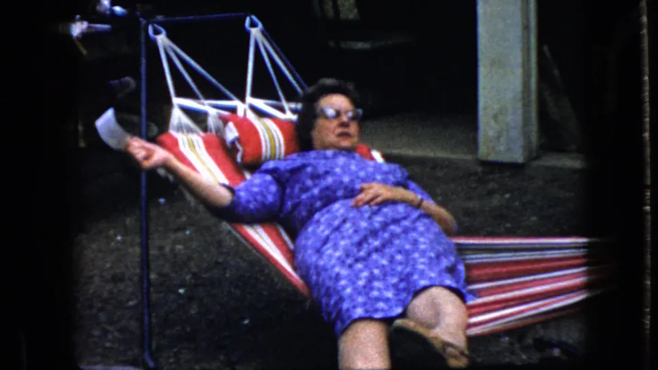 A woman wearing sunglasses and a purple patterned dress reclines in a red, white, and blue striped hammock in the 1969 California home-movie footage.