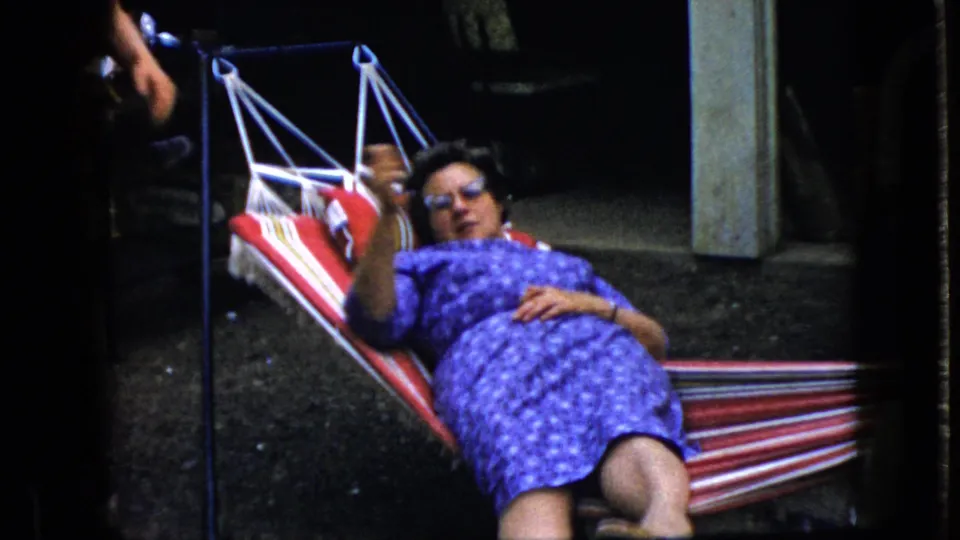 The frame shows a woman in sunglasses reclining in a striped hammock, raising one arm as the handheld 8mm camera frames her from close range outdoors.