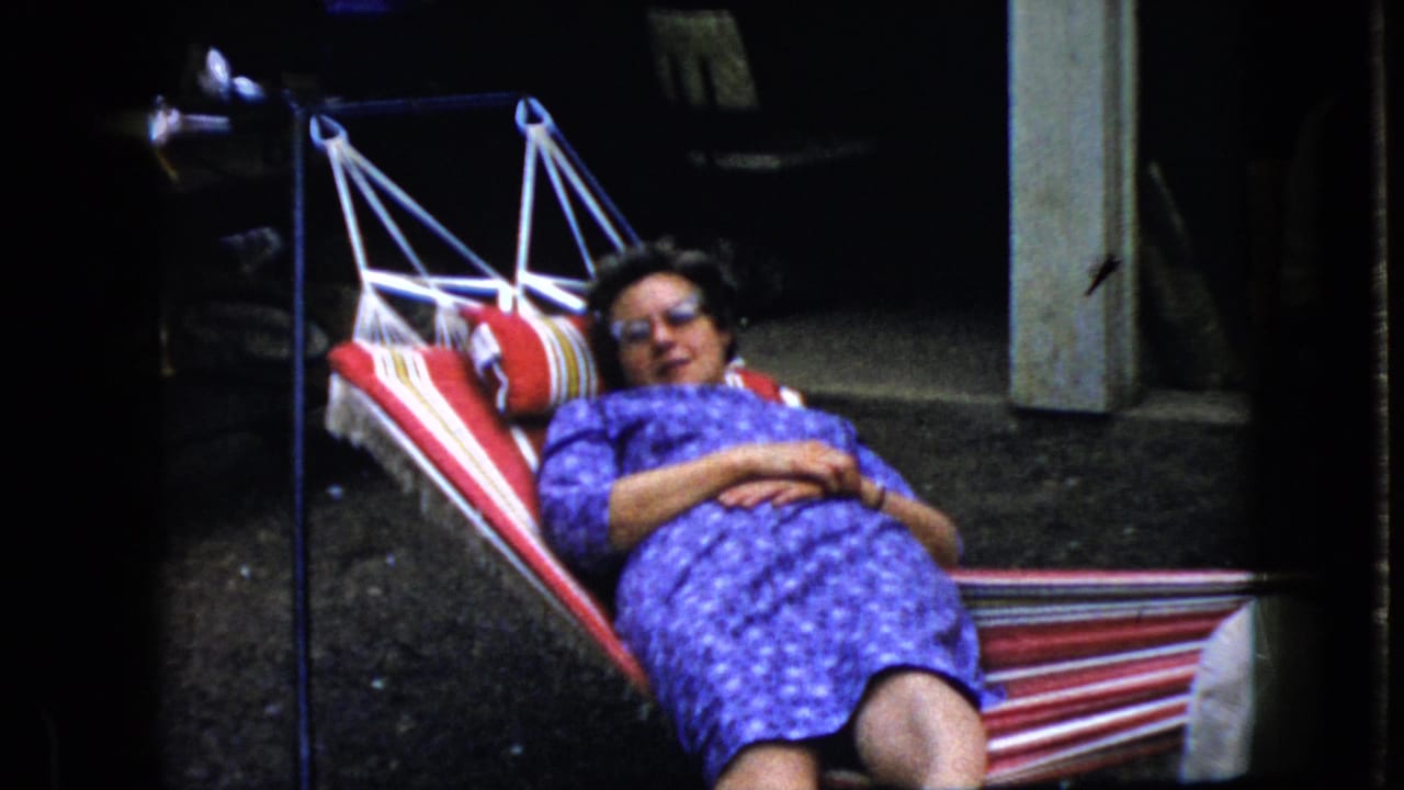 Adult woman in sunglasses and a purple patterned dress reclines in a red-and-white striped hammock in California home-movie footage from 1969.