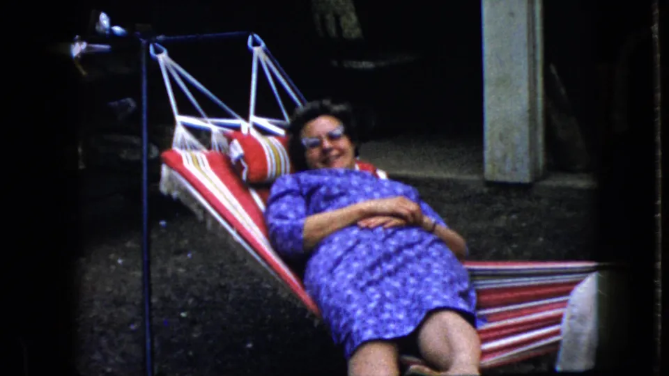 The woman smiles toward the camera while lying back in the striped hammock, with a dark outdoor background and pale post behind her.