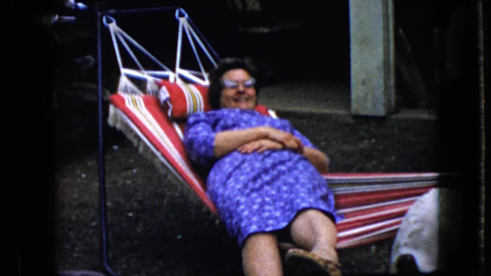 Adult woman in a purple patterned dress rests with arms folded in a striped hammock, captured with saturated 8mm color and soft home-movie focus.