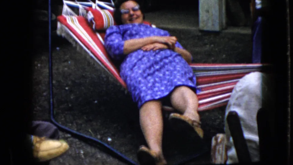 The woman continues reclining in the striped hammock as partial foreground legs and shoes appear near the lower edge of the 1969 California home-movie frame.