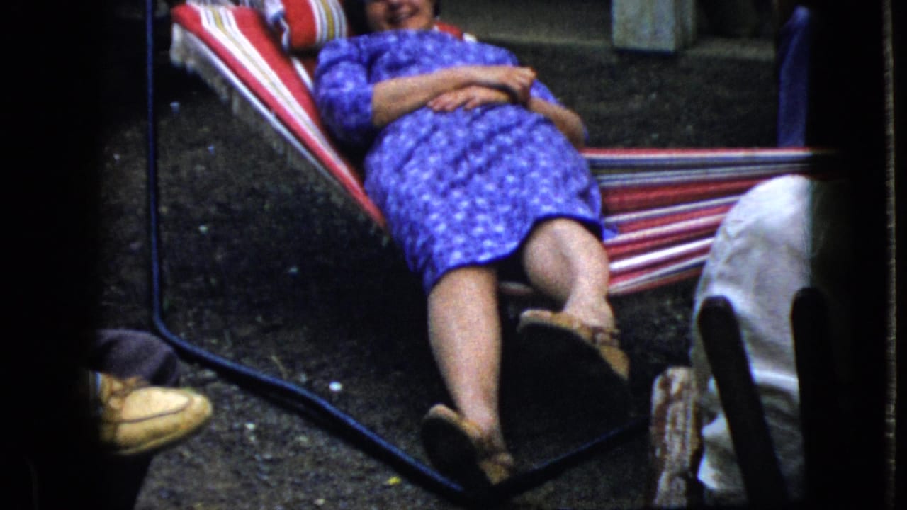 In this 1969 California home-movie frame, a woman in a blue patterned dress reclines in a red-and-white striped hammock with a pillow behind her and partial legs nearby.