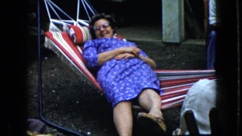 A 1969 California home-movie frame shows a smiling woman in glasses and a blue patterned dress resting in a red-and-white striped hammock in a yard.