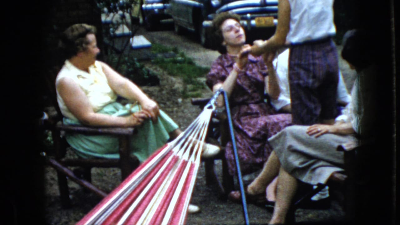 In 1969 California home-movie footage, several women gather outdoors near a red-and-white striped hammock, with chairs, shrubs, and parked mid-century cars behind them.
