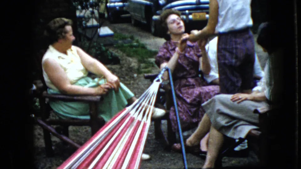 In 1969 California home-movie footage, several women gather outdoors near a red-and-white striped hammock, with chairs, shrubs, and parked mid-century cars behind them.