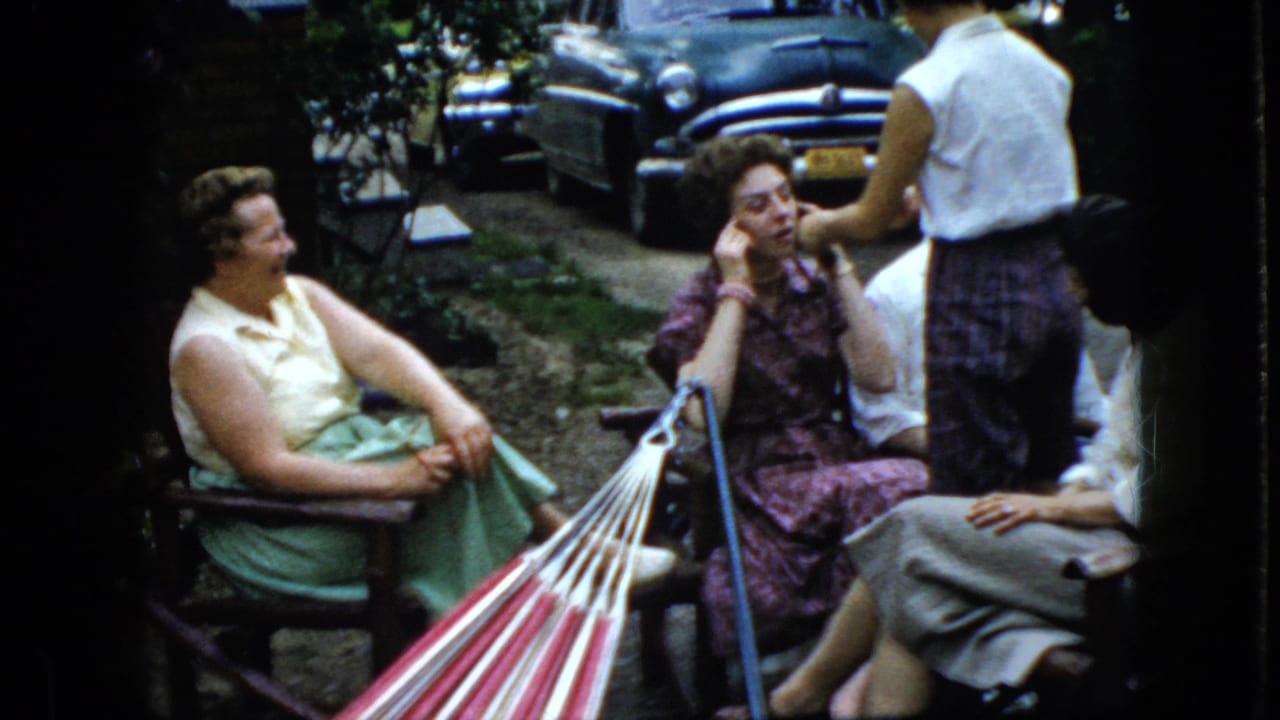 Women talk beside a hammock while a standing figure faces them near parked cars.