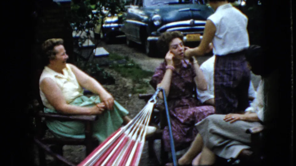 A group of women converse in an outdoor seating area beside a striped hammock, with one standing figure partly turned away and older cars visible beyond the garden path.