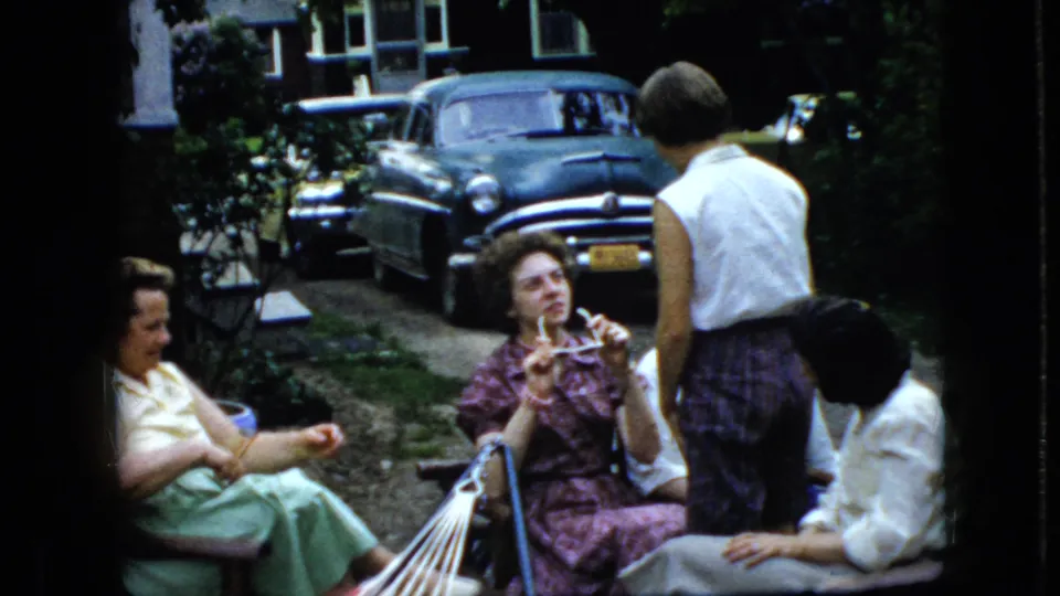 A woman in a patterned dress gestures with her hands as other women sit around a striped hammock in a California backyard scene from 1969.