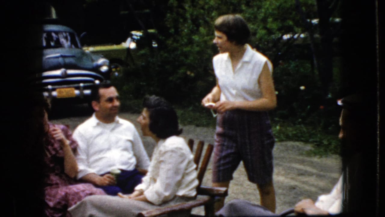 Three adults sit outdoors while a woman in a sleeveless blouse stands nearby, with an older car behind them.