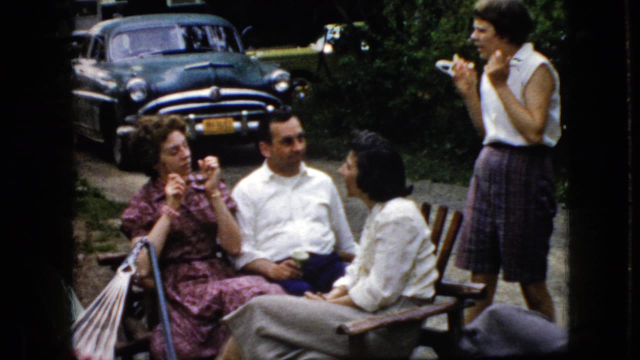 Four adults gather outdoors, with one standing woman gesturing and three people seated near a parked older car.