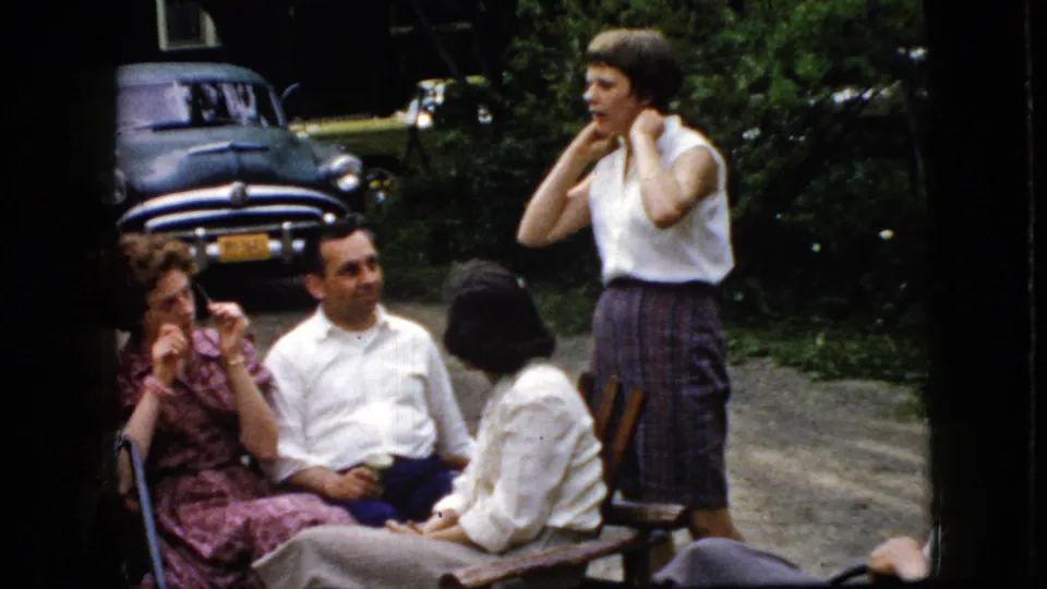 A woman in a white sleeveless blouse stands beside seated adults in a 1969 California home-movie scene, with an older parked car visible behind the backyard gathering.