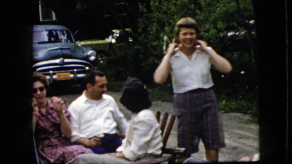 A standing woman smiles toward the camera during a backyard gathering, while three adults remain seated and a dark older car is parked behind them in the 1969 California home-movie footage.