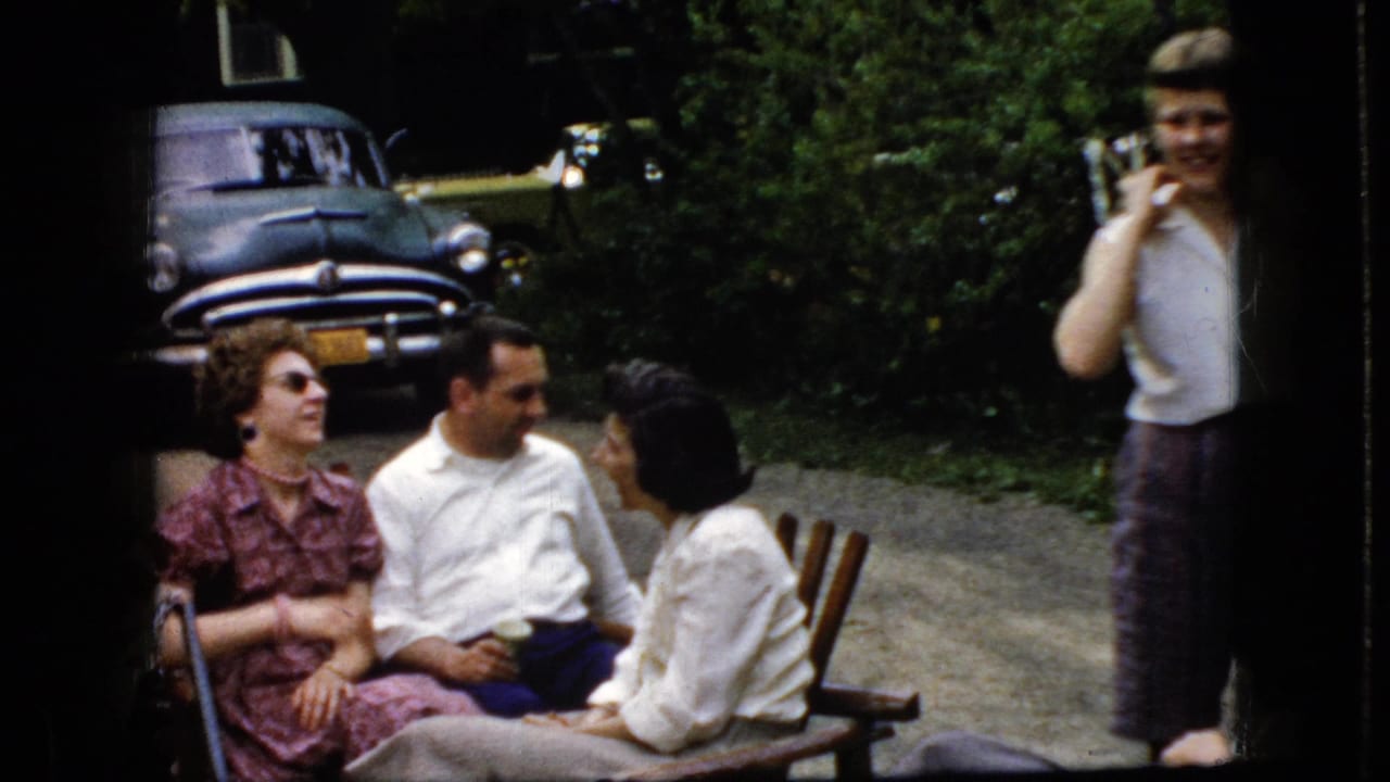 Adults sit and stand near a wooden bench and a dark classic car in a wooded outdoor area.