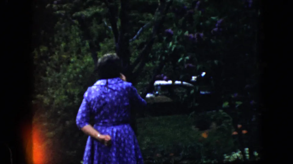 1969 California 8mm frame showing a woman in a blue patterned dress walking away through a shaded garden, with part of a dark car visible through foliage.
