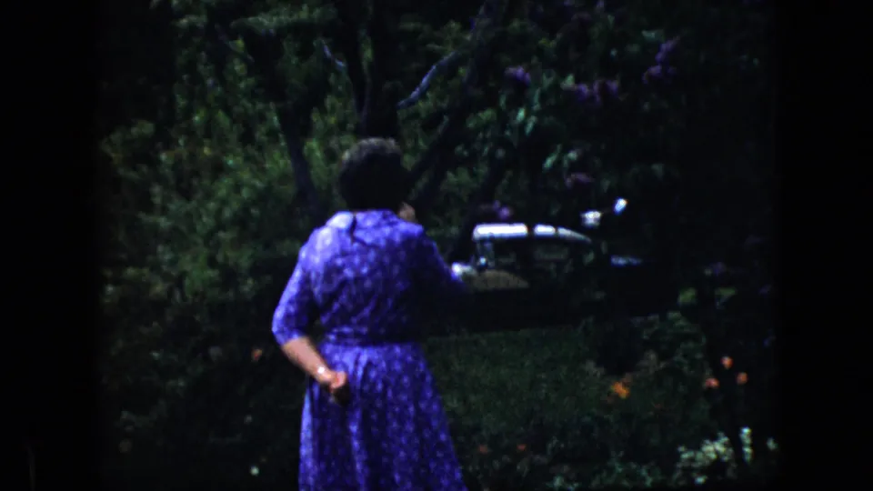 1969 California 8mm frame of a woman in a blue patterned dress seen from behind in a leafy garden, with a dark classic car partially visible beyond the plants.