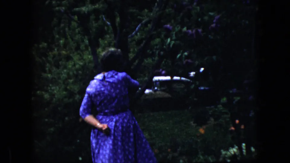 In metadata-supported 1969 California home-movie footage, a woman in a purple patterned dress walks away through dense garden foliage with a vehicle partly visible beyond the trees.