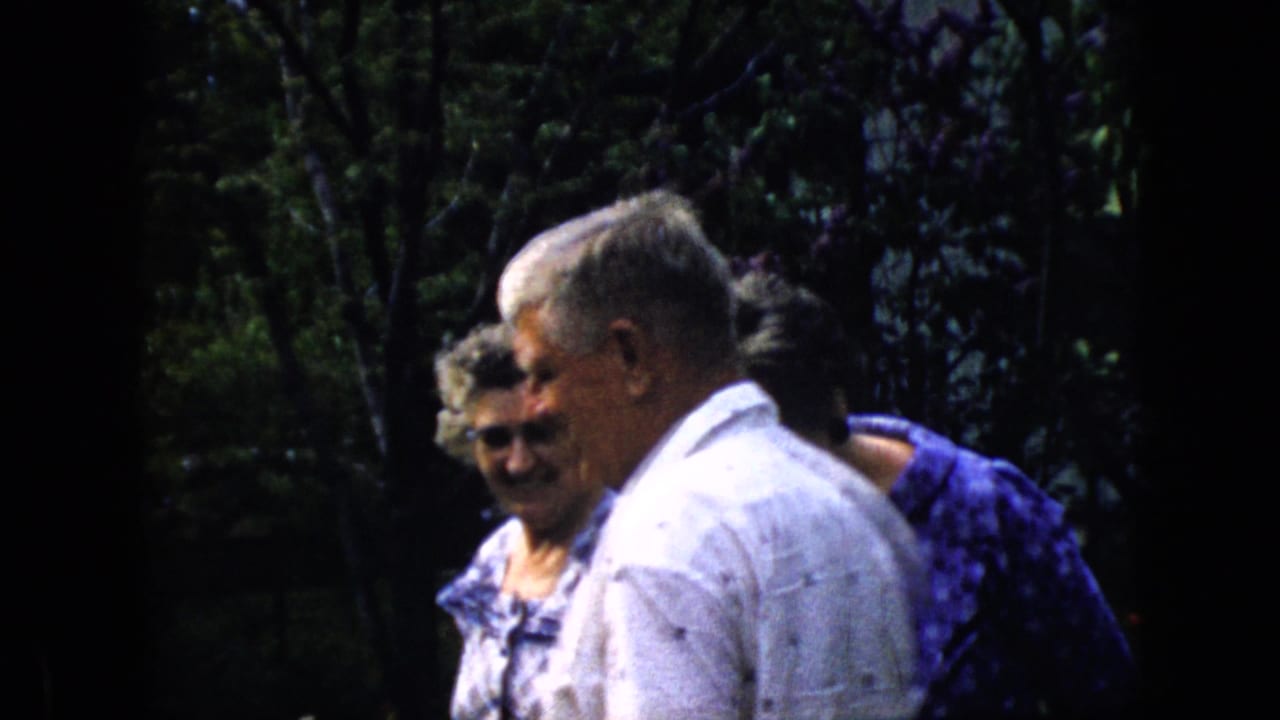 Older man in a white shirt walking beside two women in a garden.