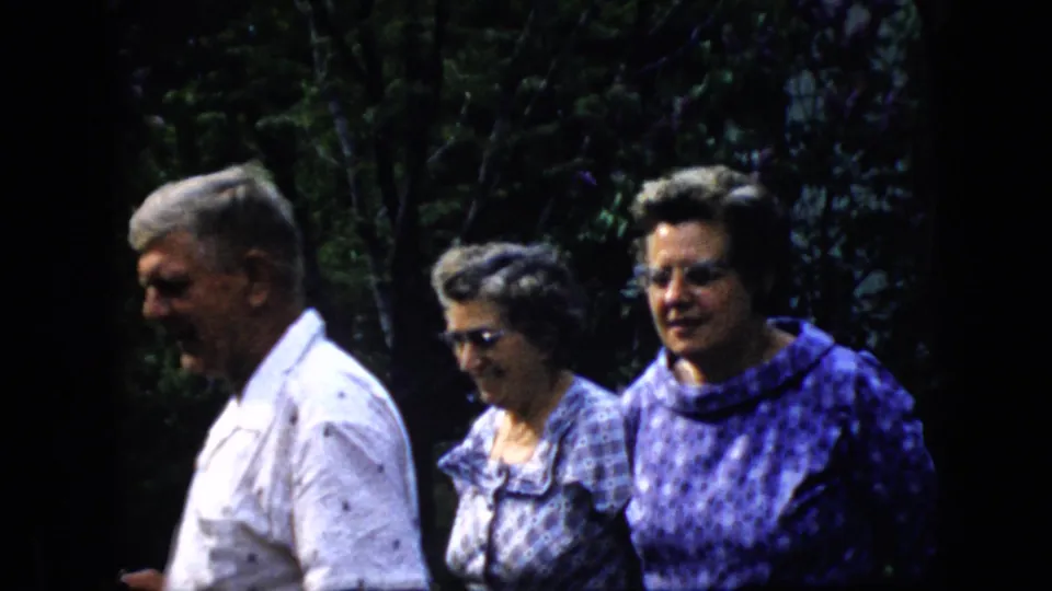 Three older adults, including a man in a white shirt and two women in patterned clothing, move together through a leafy garden area in metadata-supported 1969 California footage.