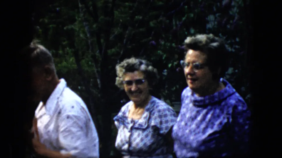 Two older women smile toward the camera in a shaded garden while an older man is partially visible at the edge of the frame in the 1969 California home-movie scene.