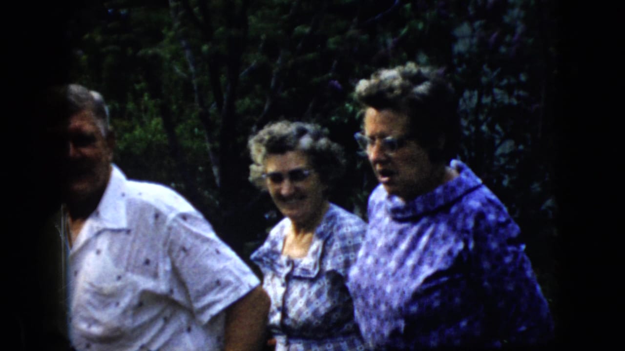 Three adults stand outdoors in a shaded wooded area as an older man in a white shirt faces the camera.