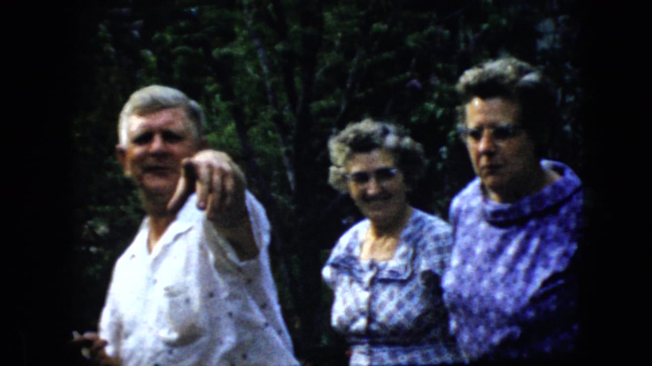 Older man points toward the camera while two women stand beside him in a wooded area.