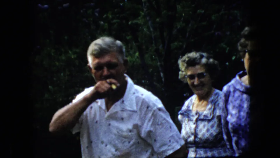A candid 1969 home-movie frame shows the older man with his hand near his mouth, with two women beside him against a dark wooded background.