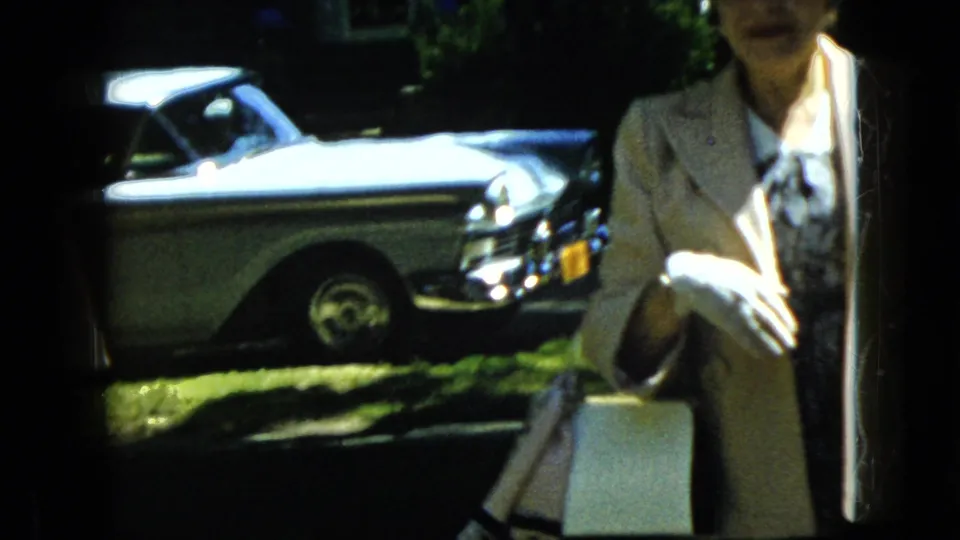 A brief 1969 California home-movie shot shows a parked mid-century car in the background with a woman partly visible at the right edge of the frame.
