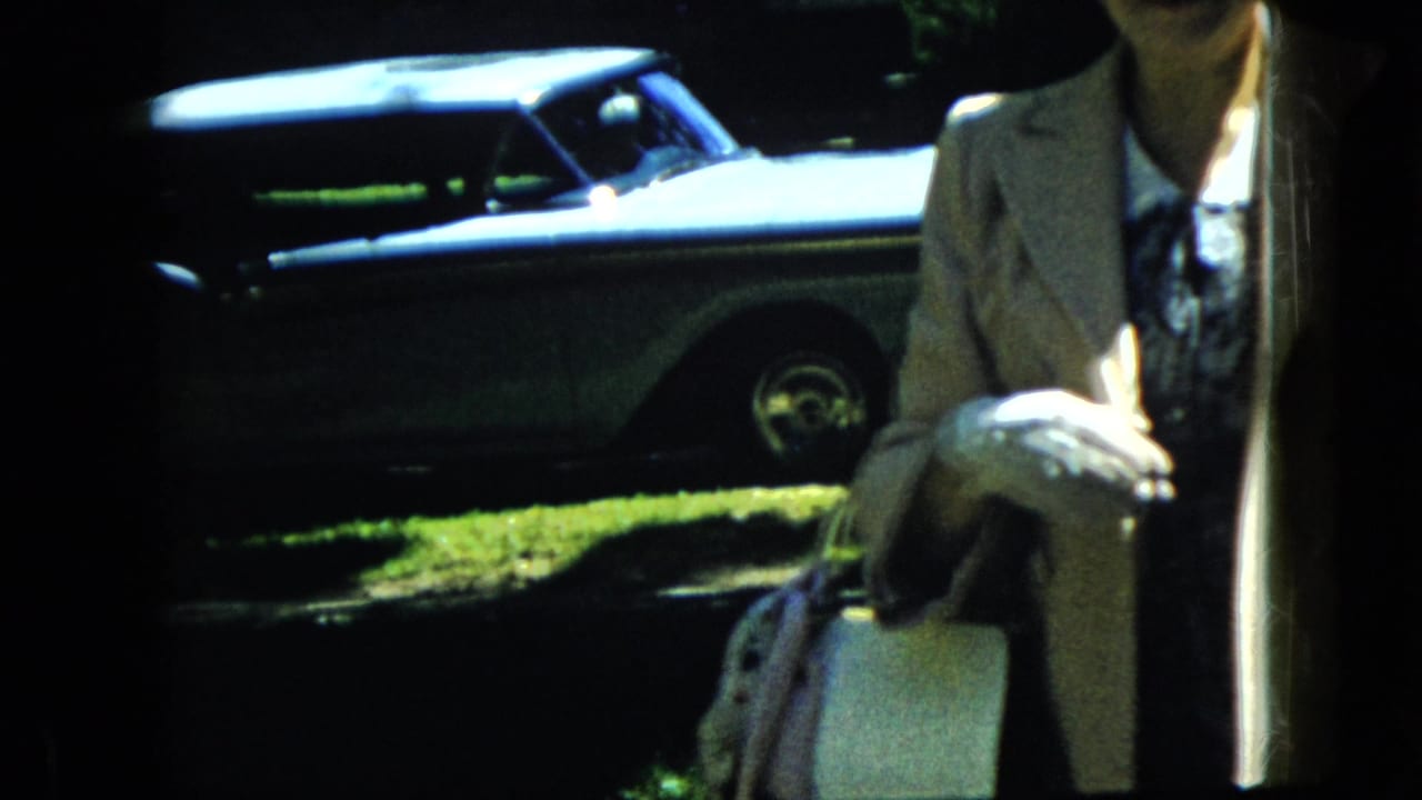 In a 1969 California home-movie frame, a woman in a light-colored suit holds white gloves and a bag while standing in a sunny yard near a parked sedan.