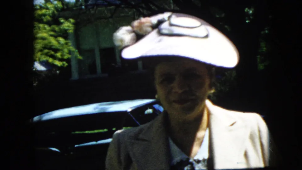 A woman wearing a wide-brimmed floral hat and light suit smiles toward the camera in a sunny California yard, with a dark sedan and house exterior behind her.