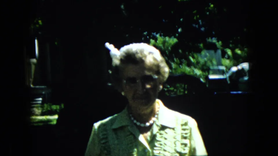 An older woman wearing glasses, a pearl necklace, and a patterned dress stands in strong sunlight and shade during a 1969 California home-movie moment.