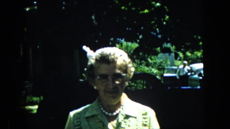 A woman in eyeglasses, pearls, and a patterned dress faces the home-movie camera in a shaded outdoor yard with foliage and parked cars in the background.