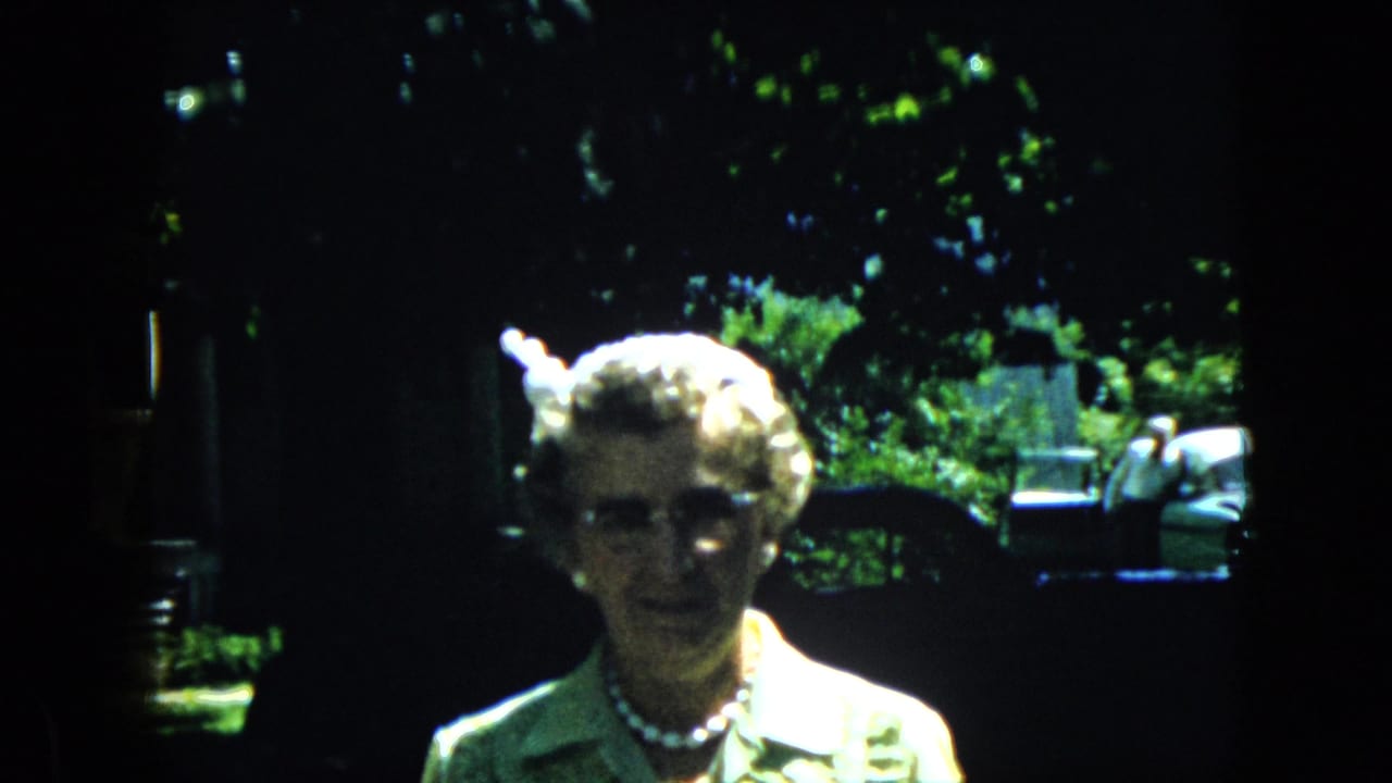 Elderly woman in a light collared top, glasses, and pearl necklace faces the camera in a California backyard, consistent with the supplied 1969 home-movie metadata.