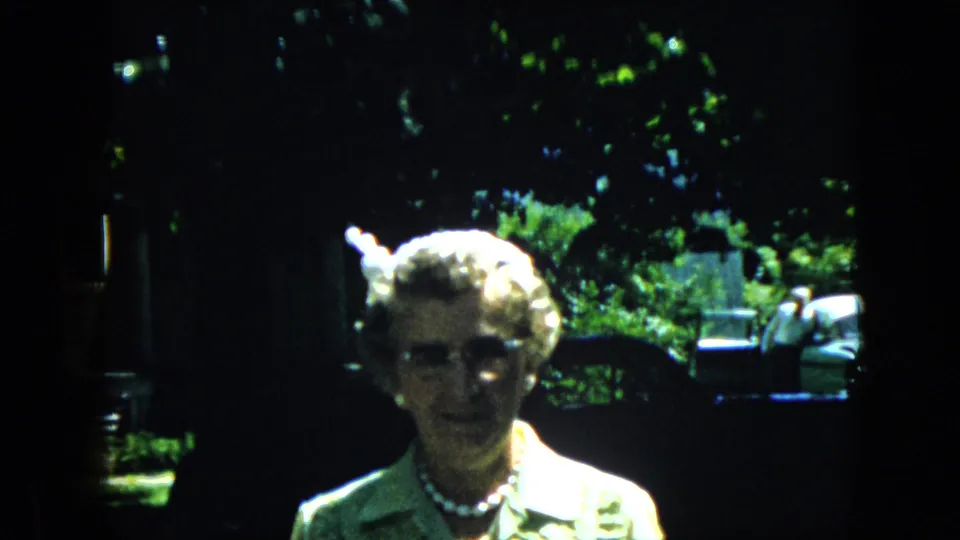 Elderly woman in a light collared top, glasses, and pearl necklace faces the camera in a California backyard, consistent with the supplied 1969 home-movie metadata.