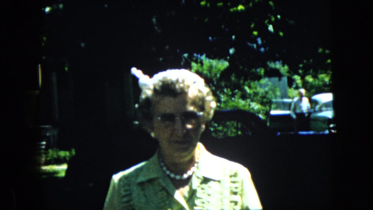 Elderly woman remains facing the camera with trees and a parked car behind her.