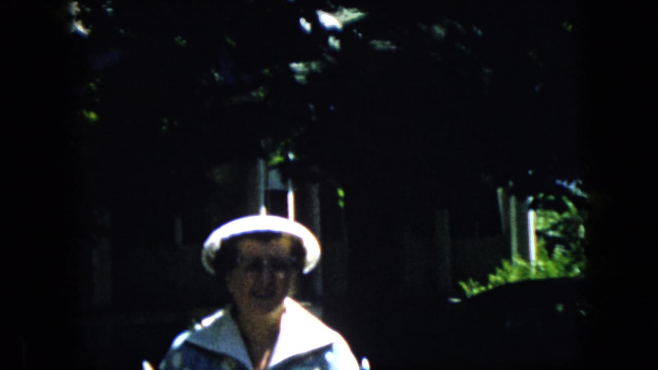 Older woman in a white hat and glasses walks toward the camera in a shaded garden.