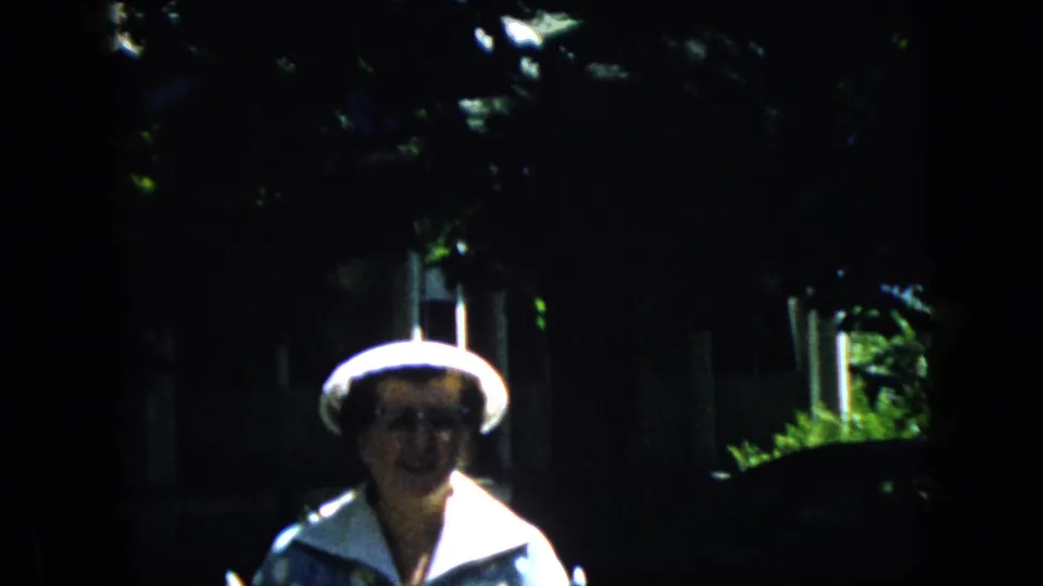 In this 1969 California home-movie frame, an older woman wearing a white hat, glasses, and a light collared jacket approaches the camera in a dark, leafy residential garden.